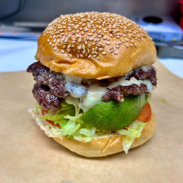 A double cheeseburger with melted cheese, lettuce, tomato, and avocado on a sesame seed bun.