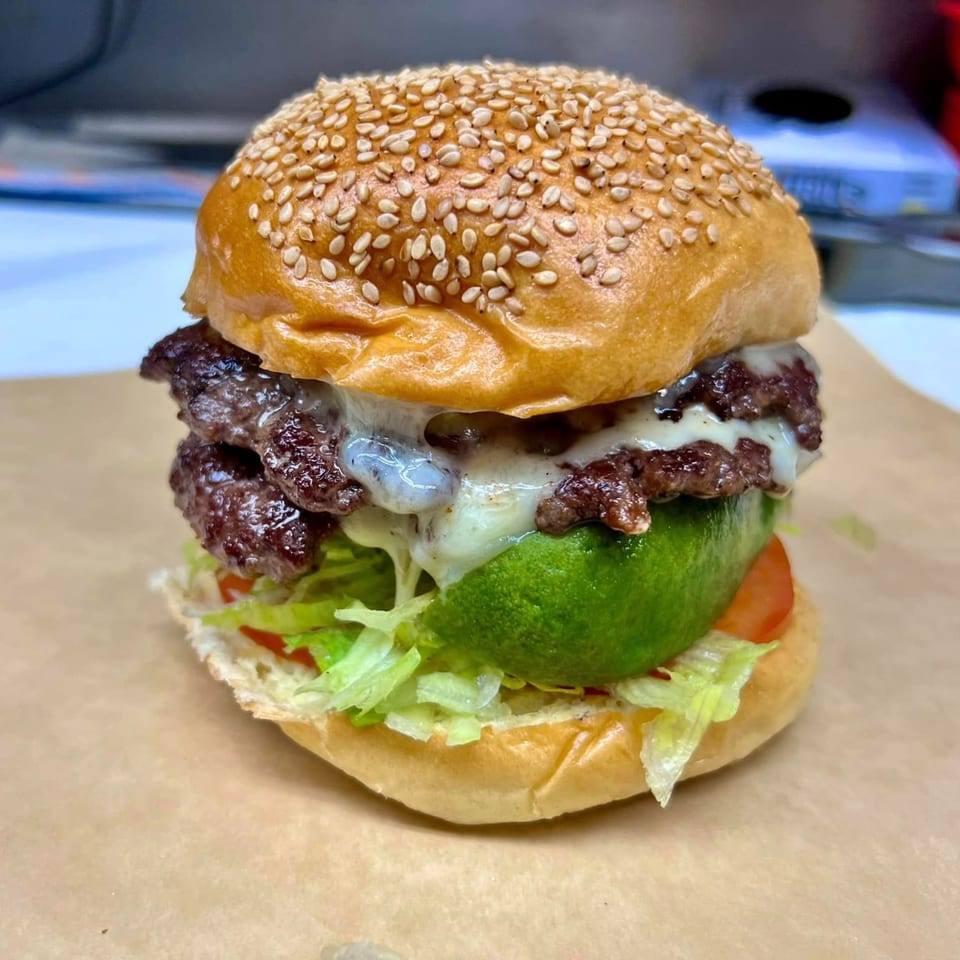 A double cheeseburger with melted cheese, lettuce, tomato, and avocado on a sesame seed bun.