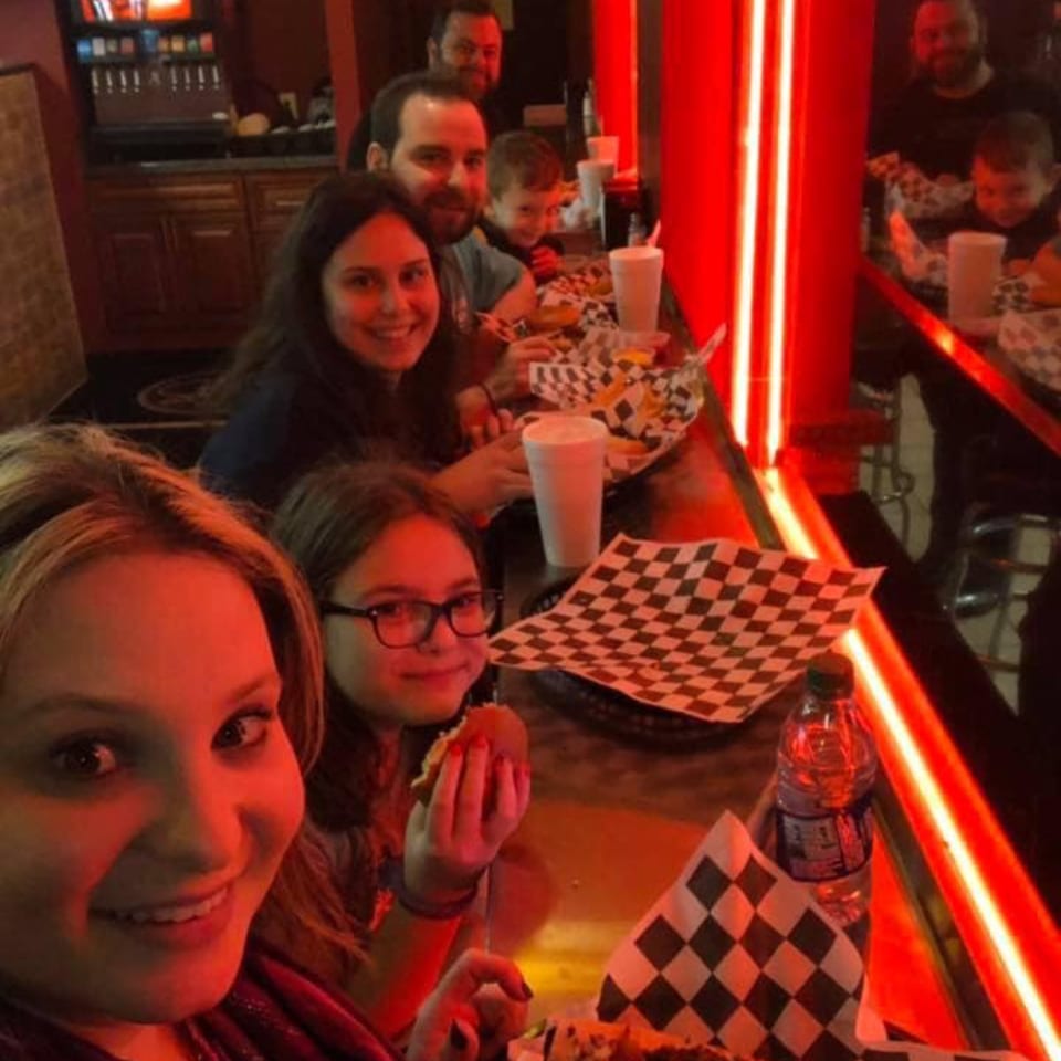 A family of five, including children, enjoys cheesesteaks and fries at a counter with red lighting.